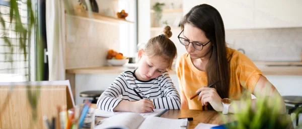 Eine Mutter sitzt mit ihrer Tochter zuhause am Küchentisch, das Kind schreibt etwas.