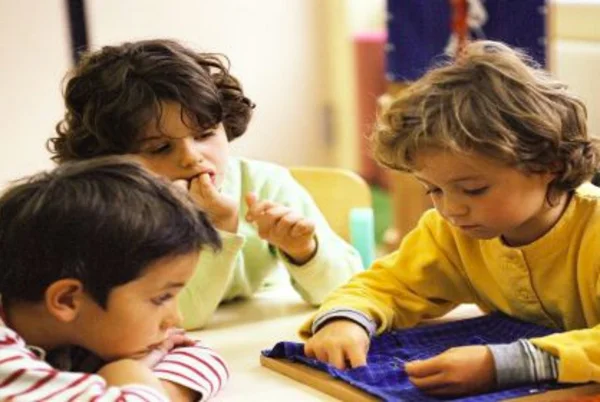 Drei Kinder sitzen am Tisch bei der Handarbeit an einem Stück Stoff. / El işçiliği de çocuklar