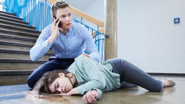 Erste Hilfe Ein Ersthelfer ruft per Telefon um Hilfe während er neben einer bewusstlos auf dem Boden liegenden Person kniet.