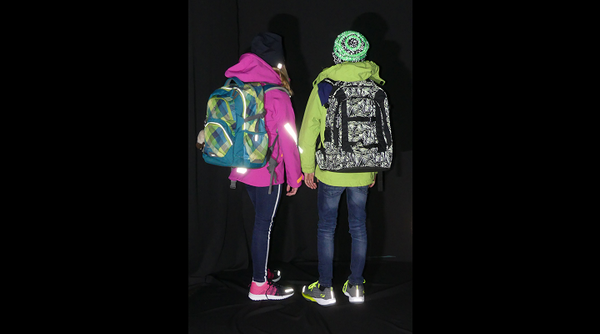 Zwei Kinder in heller Kleidung und mit Rucksack in Dunkelheit auf dem Weg zur Schule