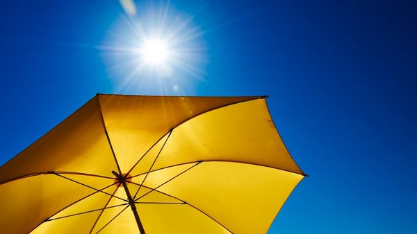 Ein gelber Sonnenschirm vor einem strahlend blauen Himmel mit Sonne.