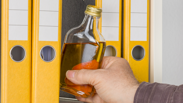 Eine Flasche Alkohol wird von einer Hand zwischen Aktenordnern aus einem Regal herausgenommen.